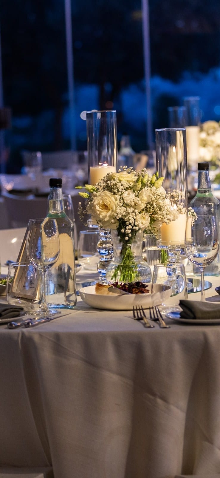 A round table laid for a reception at dusk: white linen, candles and a bouquet of roses, with the city lights beyond the windows.